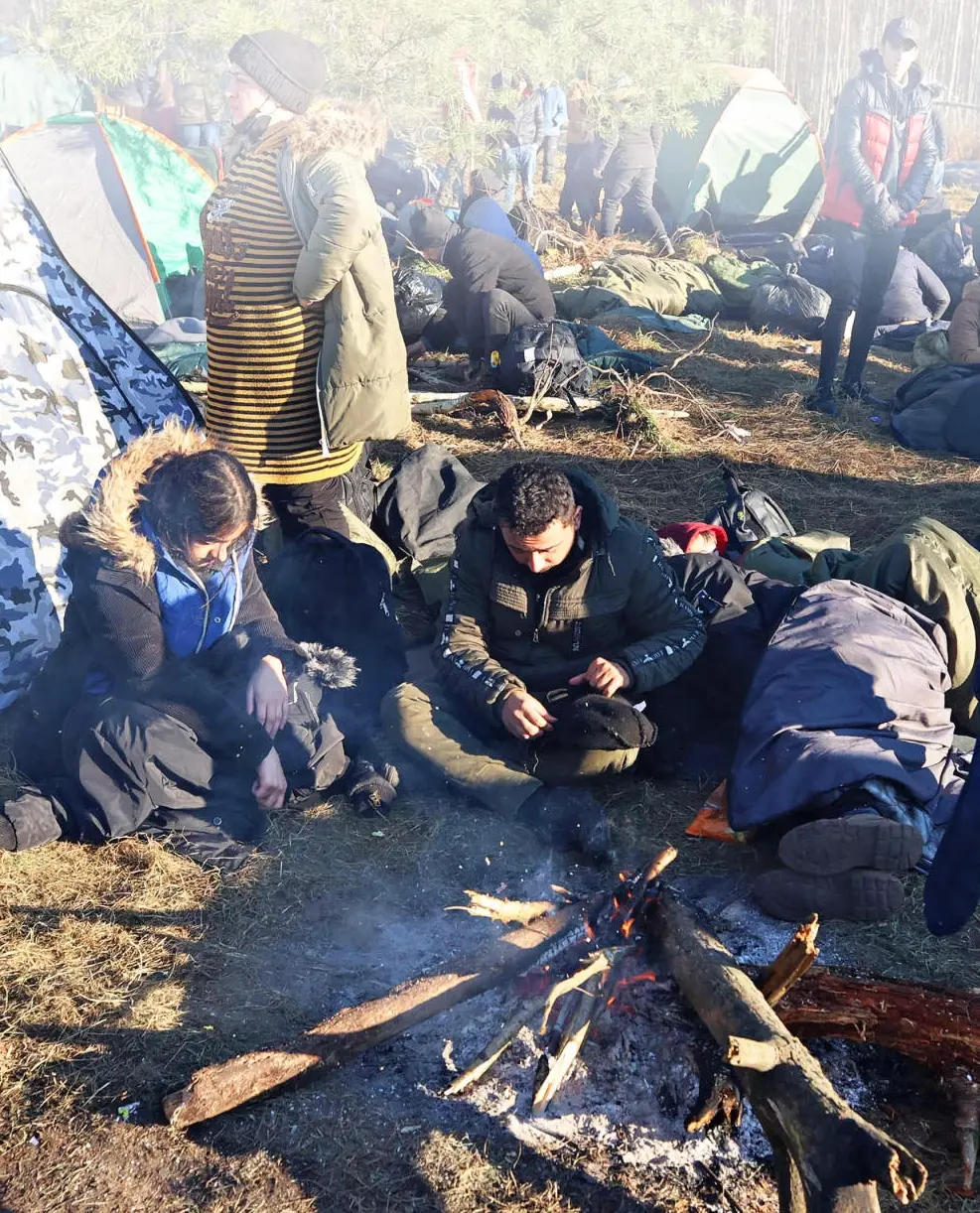 Menschen in Winterkleidung sitzen auf dem Waldboden vor Zelten und um ein Feuer.