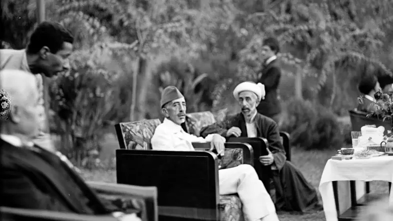 Zu sehen ist eine Schwaz-Weiß-Aufnahme des früheren Königs des Irak, Feisal I., und seinem Bruder Abdullah, dem Emir und König von (Trans-)Jordanien. Beide sitzen in einem Garten auf zwei Sesseln neben einem gedeckten Tisch und weiteren Gästen. 