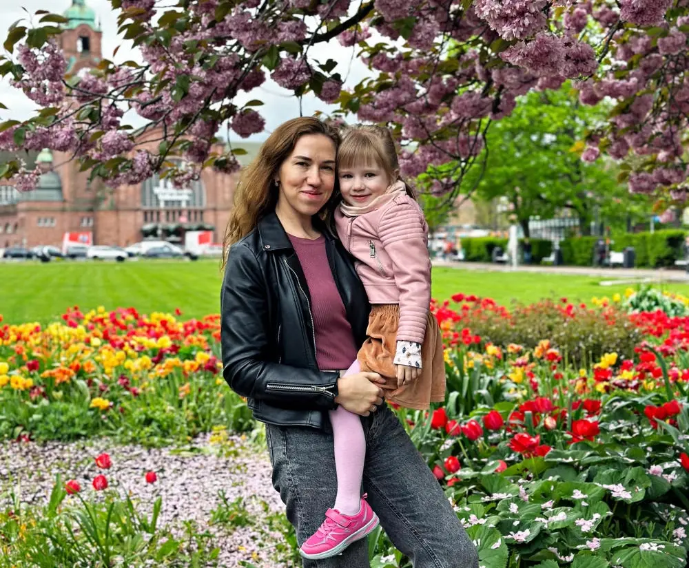 Olha hat ihre Tochter auf dem Arm, beide lächeln und stehen vor bunten Blumen