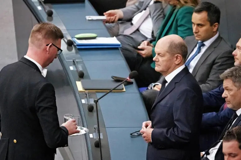 Ein Plenarassistent überreicht ein Glas mit Wasser an Bundeskanzler Olaf Scholz.