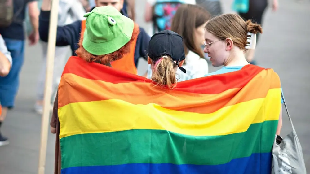 Drei Personen stehen eng beieinander und halten die Regenbogenflagge am Rücken
