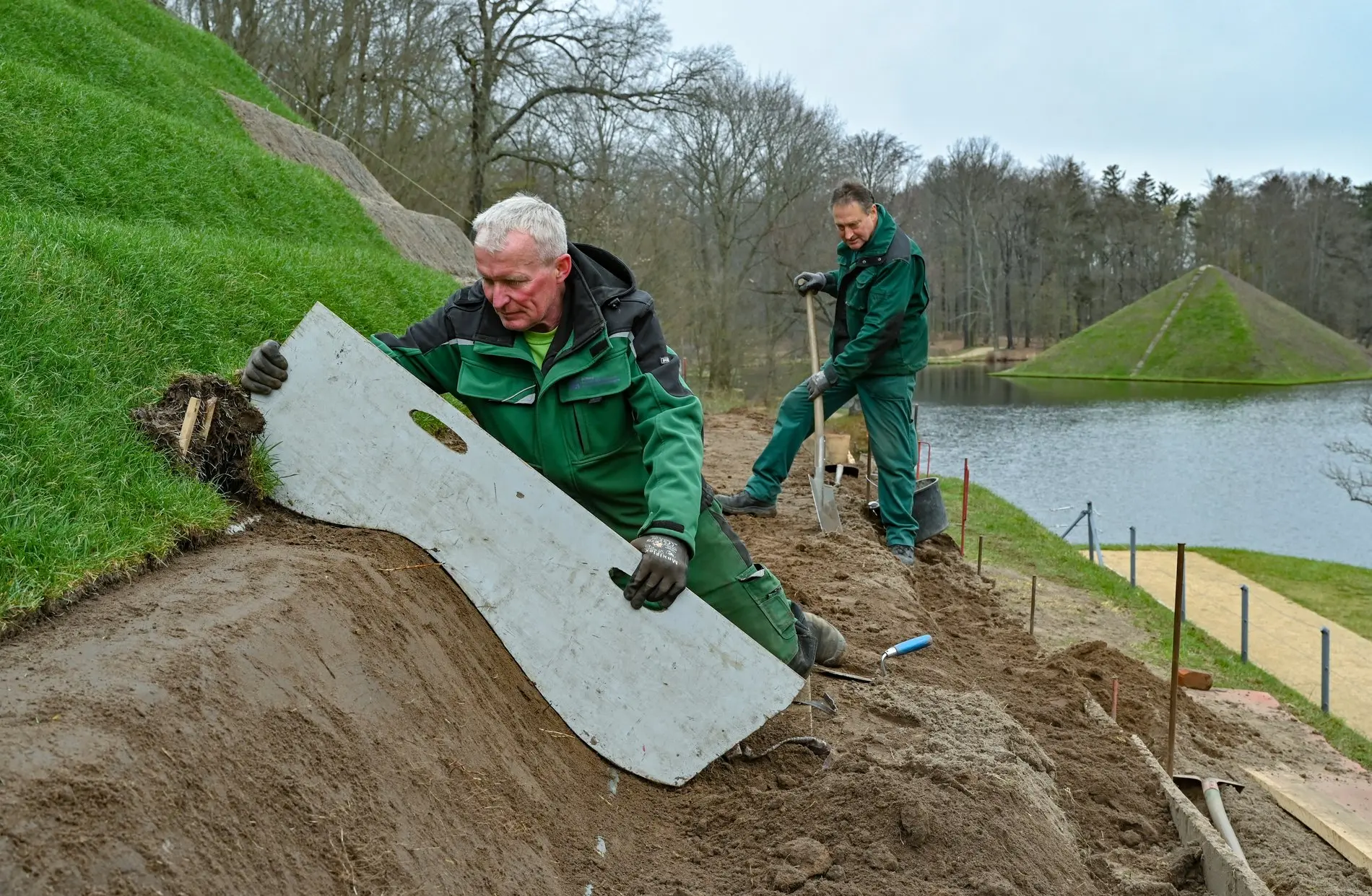 Zwei Freiwillige in Arbeitskleidung arbeiten an einem grünen Hang.