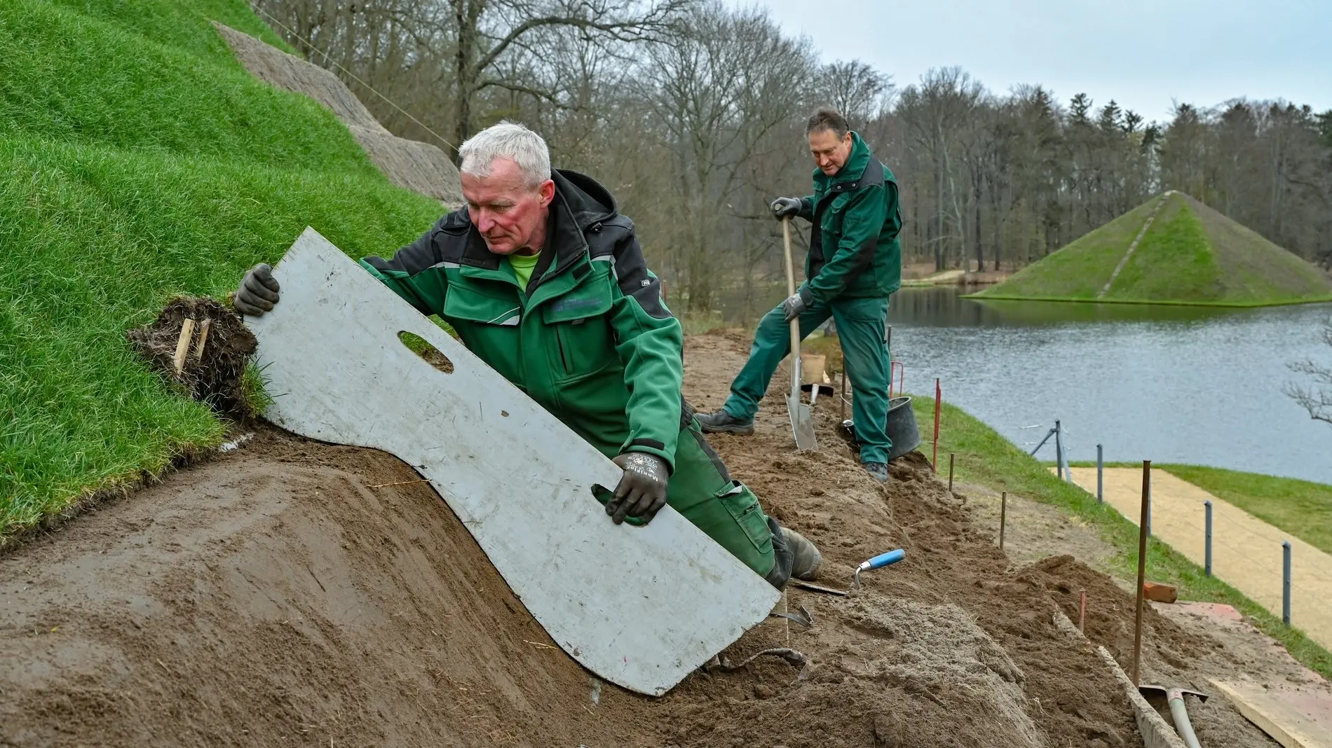 Zwei Freiwillige in Arbeitskleidung arbeiten an einem grünen Hang.