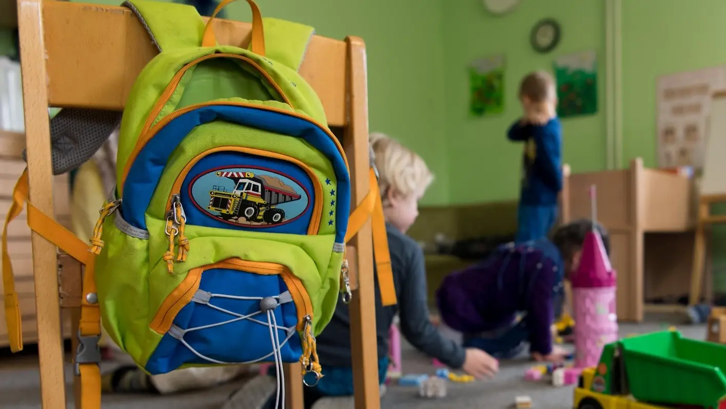 Ein grüner Kinderrucksack mit blauen und orangenen Applikationen hängt an einem Kinderholstuhl.