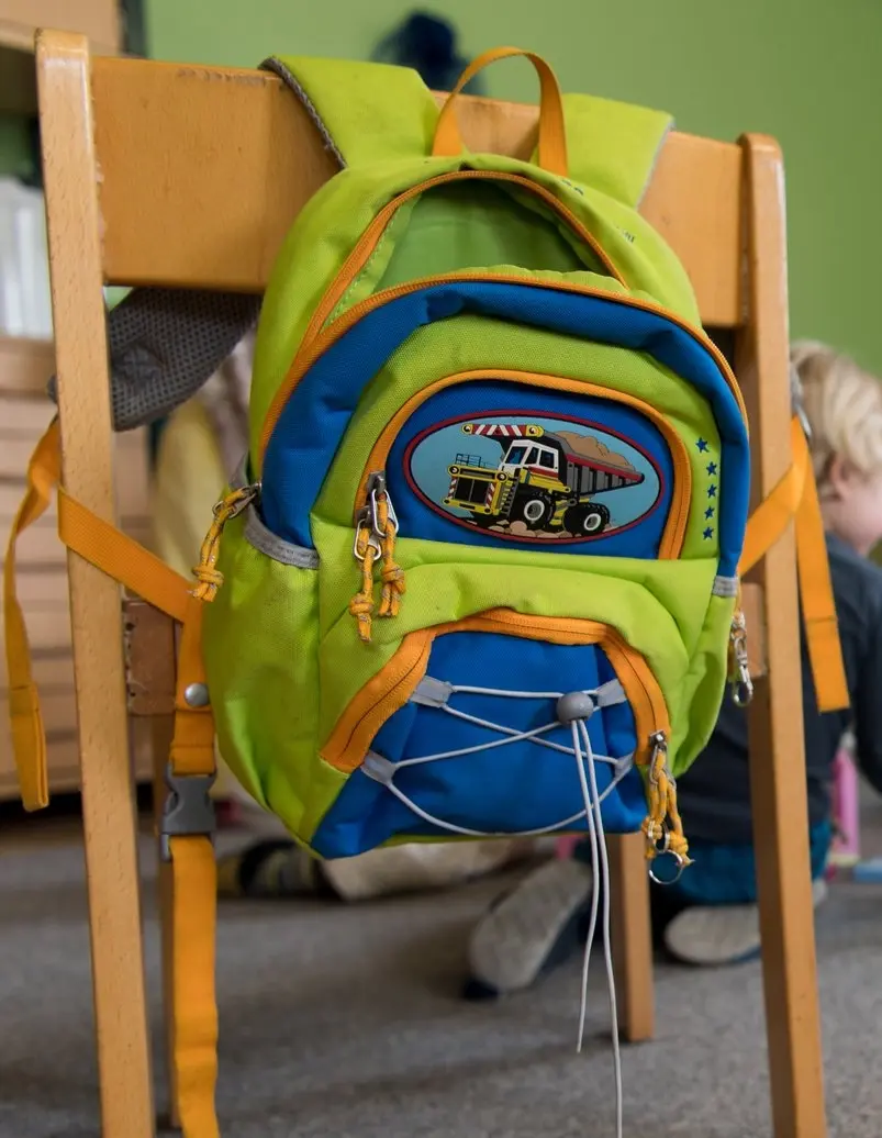 Ein grüner Kinderrucksack mit blauen und orangenen Applikationen hängt an einem Kinderholstuhl.