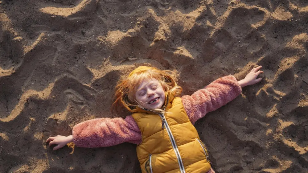 Unverzichtbare Außenseiter Kind liegt mit ausgestreckten Armen im Sand und lacht