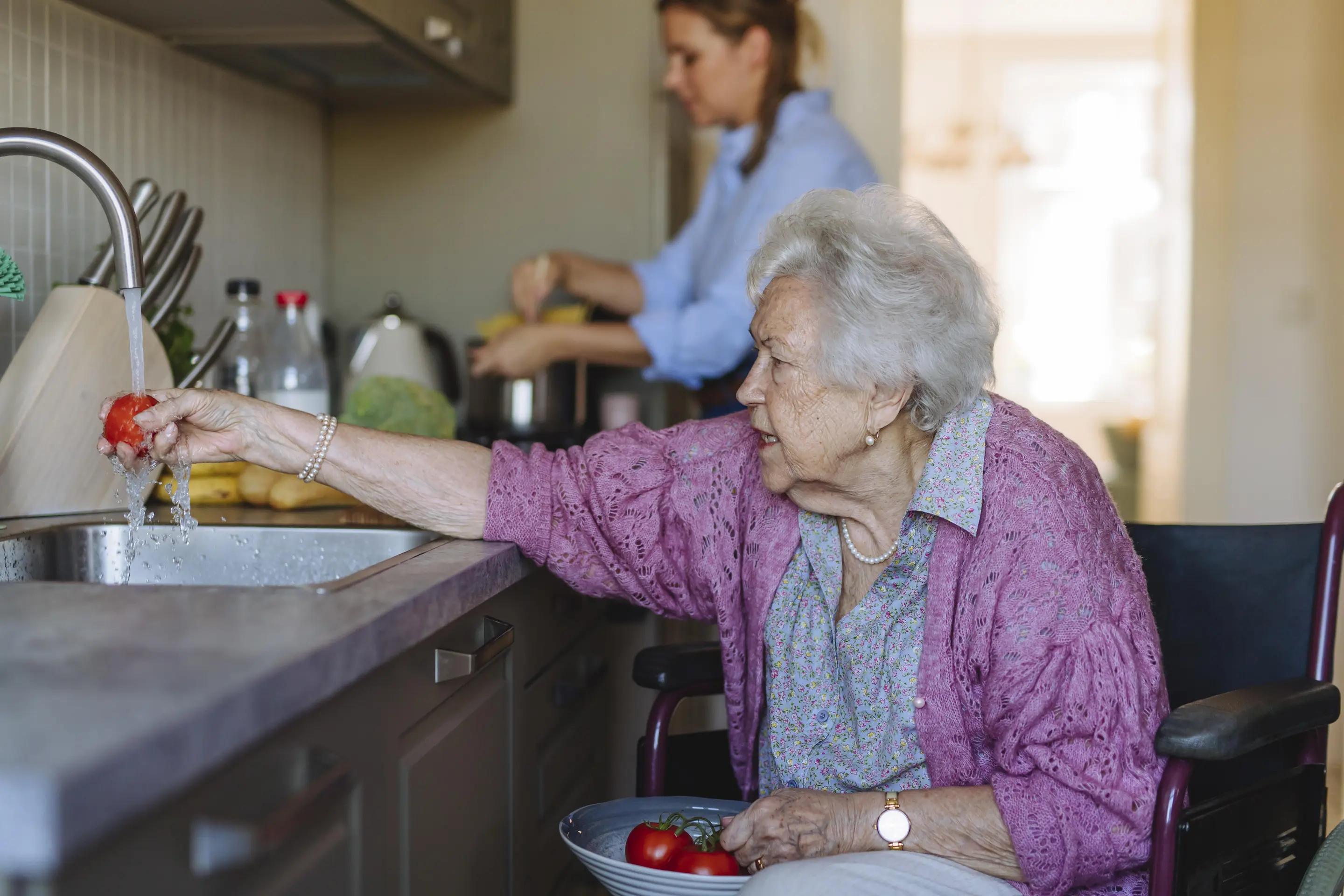 Ältere Frau wäscht Tomaten, während im Hintergrund eine Pflegekraft arbeitet.
