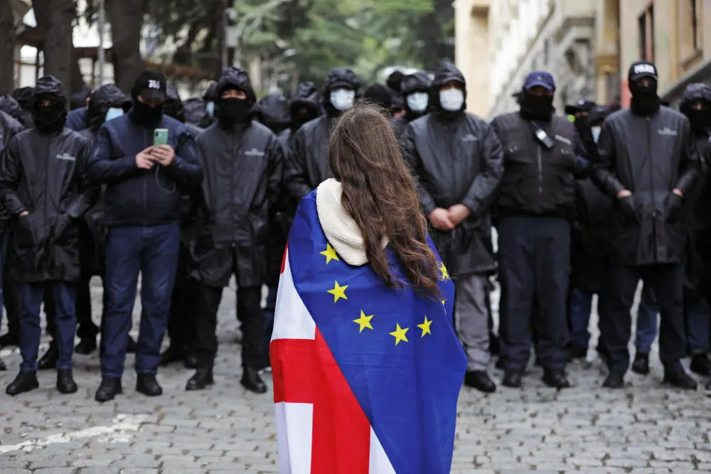 Proteste in Georgien Polizeibeamte stehen während Georgien Protesten Wache. Vor ihnen steht eine Demonstrantin mit umgehängter Flagge.