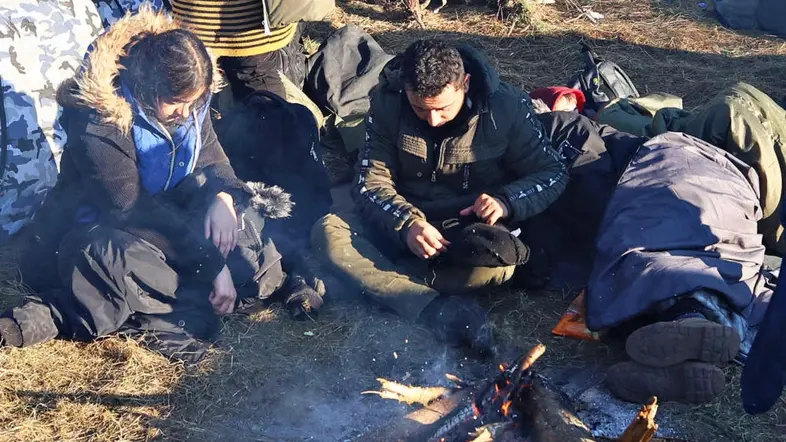 Menschen in Winterkleidung sitzen auf dem Waldboden vor Zelten und um ein Feuer.