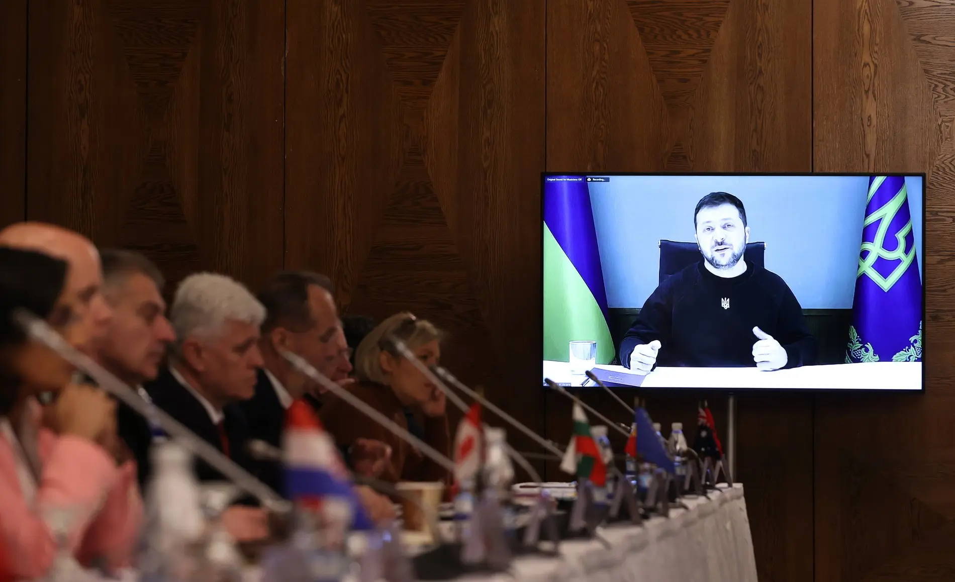 Ukrainischer Präsident Wolodymyr Selenskyj Selenskyj auf einem TV-Monitor, daneben Personen an Mikrophonen