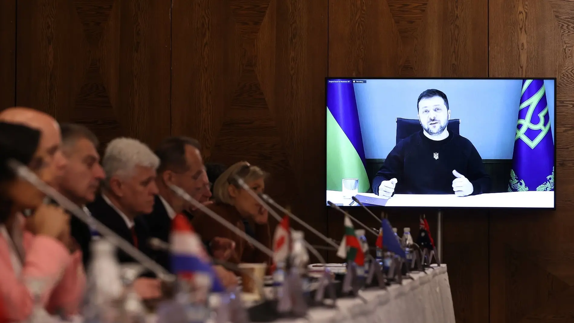 Ukrainischer Präsident Wolodymyr Selenskyj Selenskyj auf einem TV-Monitor, daneben Personen an Mikrophonen