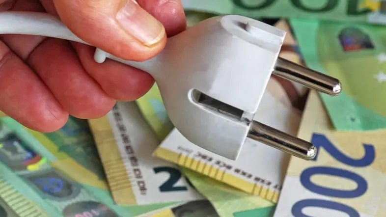 Eine Hand hält ein Stromkabel, im Hintergrund liegen 100 und 200 Euroscheine.