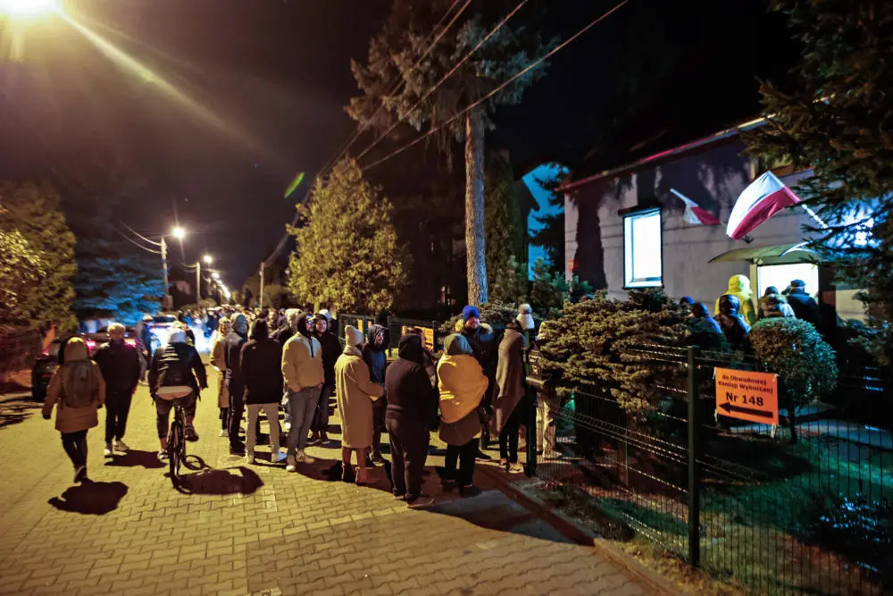 Zahlreiche Menschen stehen bei Nacht in einer langen Schlange vor einem Wahllokal mit der polnischen Flagge.
