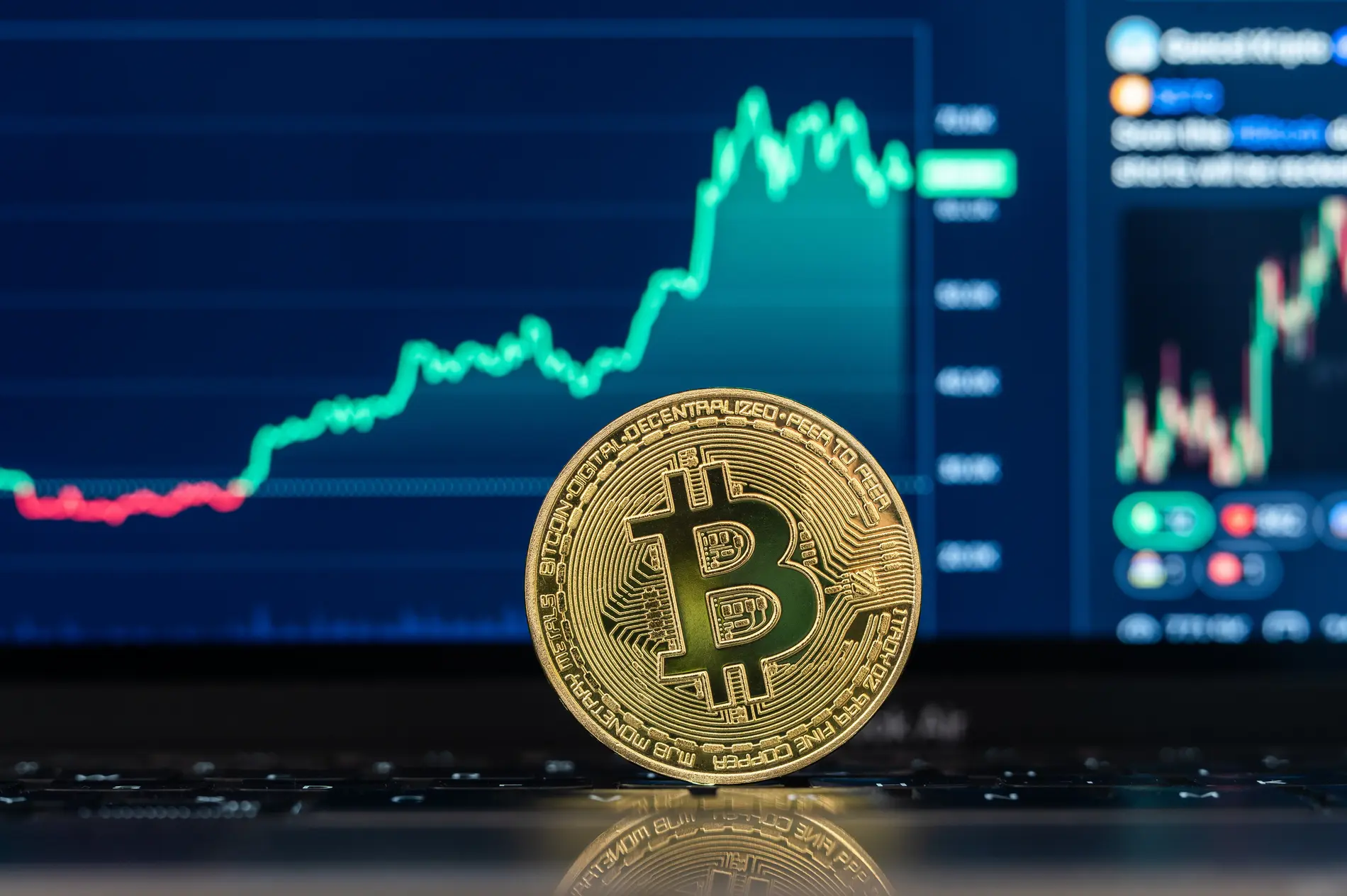 Das Logo der Kryptowährung Bitcoin (BTC) ist auf einer Münze zu sehen, die vor einem Bitcoinchart der Handelsplattform CoinMarcetCap auf einem Laptop steht.