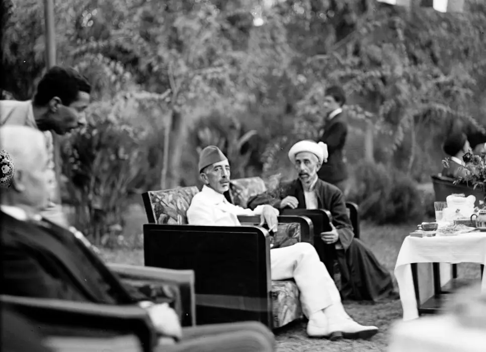 Zu sehen ist eine Schwaz-Weiß-Aufnahme des früheren Königs des Irak, Feisal I., und seinem Bruder Abdullah, dem Emir und König von (Trans-)Jordanien. Beide sitzen in einem Garten auf zwei Sesseln neben einem gedeckten Tisch und weiteren Gästen. 