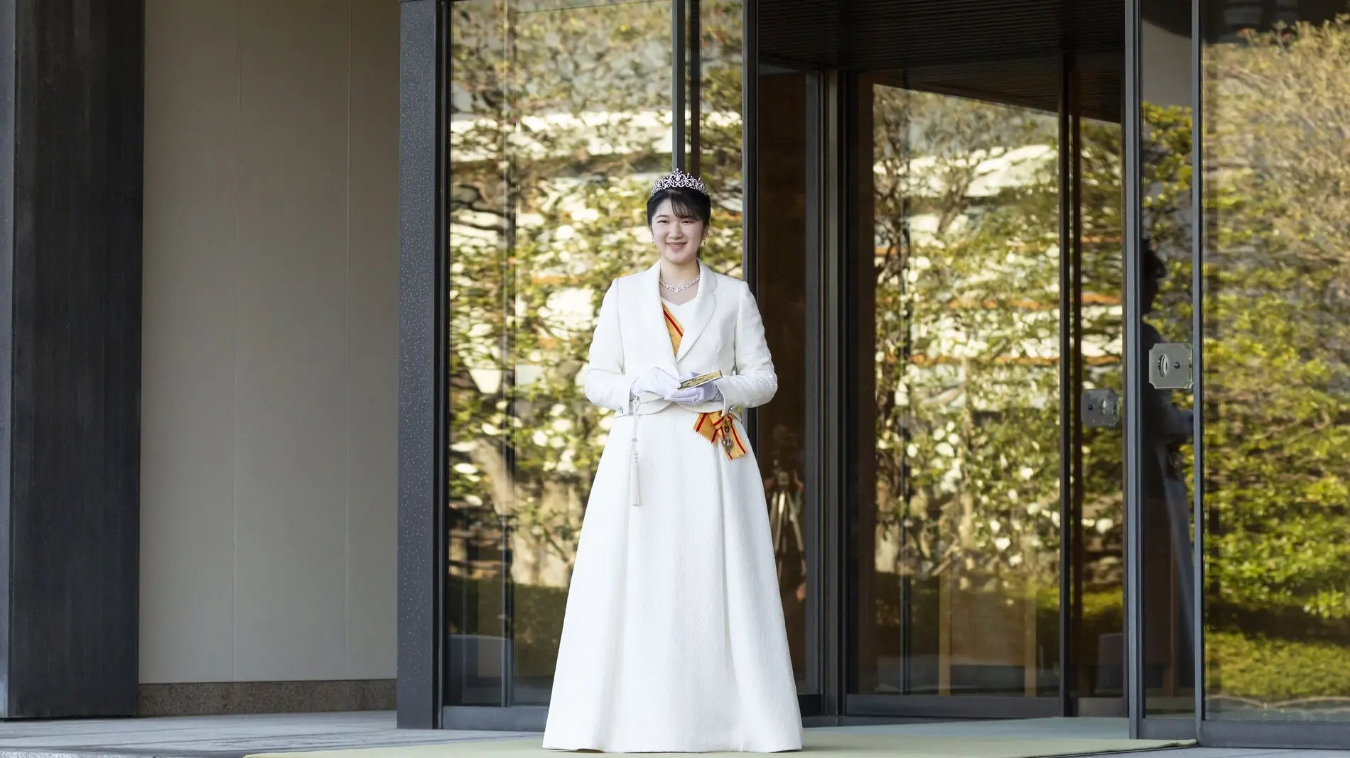 Zu sehen ist die japanische Prinzessin Aiko. Sie steht in einem weißen Kleid vor einer großen Glastür und lächelt in die Kamera. 