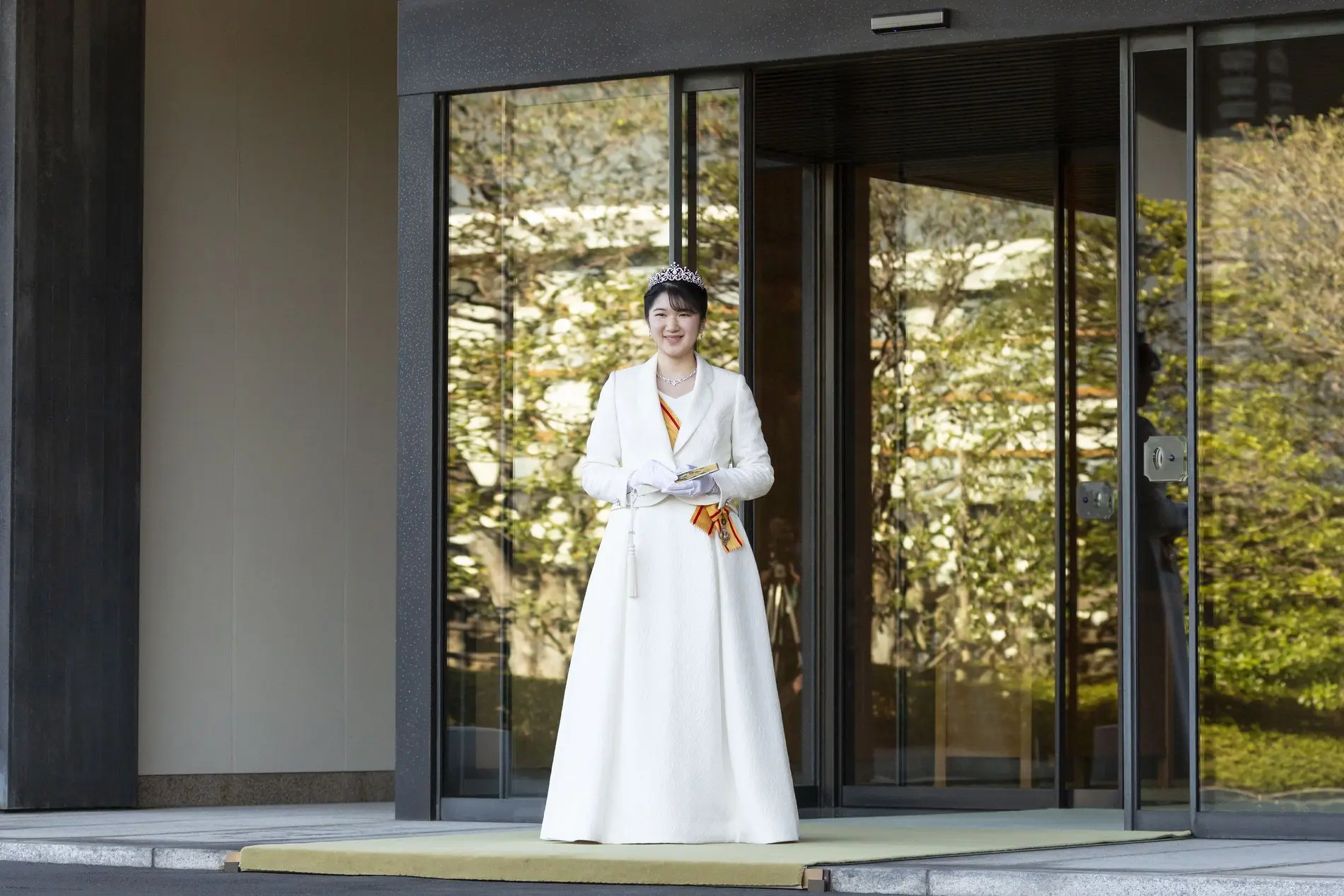 Zu sehen ist die japanische Prinzessin Aiko. Sie steht in einem weißen Kleid vor einer großen Glastür und lächelt in die Kamera.