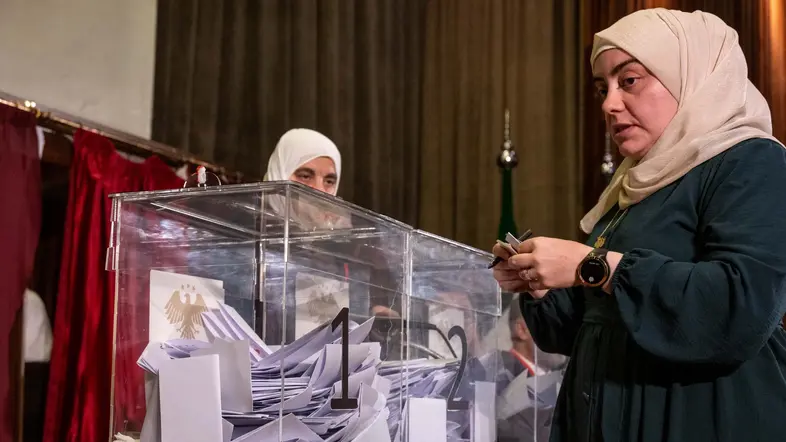 Zwei Frauen stehen an durchsichtigen Wahlurnen, in denen Wahlscheine liegen