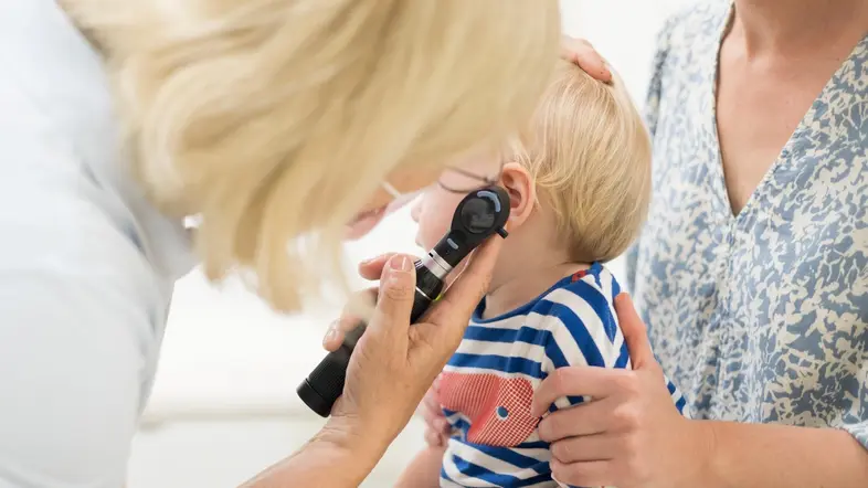 Eine Ärztin untersucht die Ohren eines kleinen Kindes, das bei seiner Mutter auf dem Schoß sitzt.