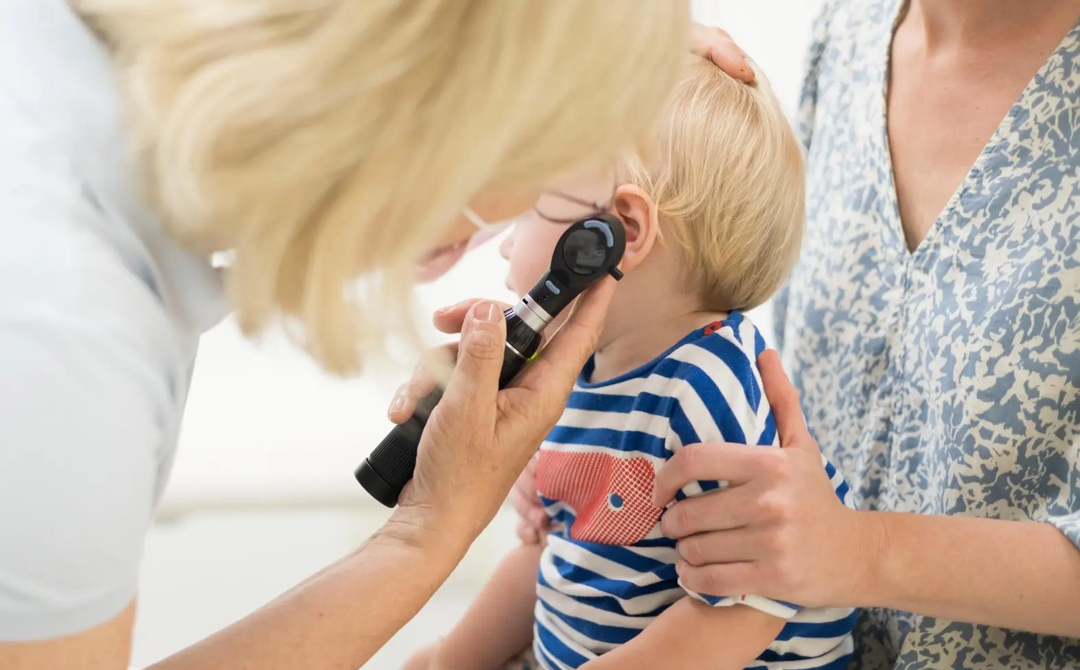 Eine Ärztin untersucht die Ohren eines kleinen Kindes, das bei seiner Mutter auf dem Schoß sitzt.