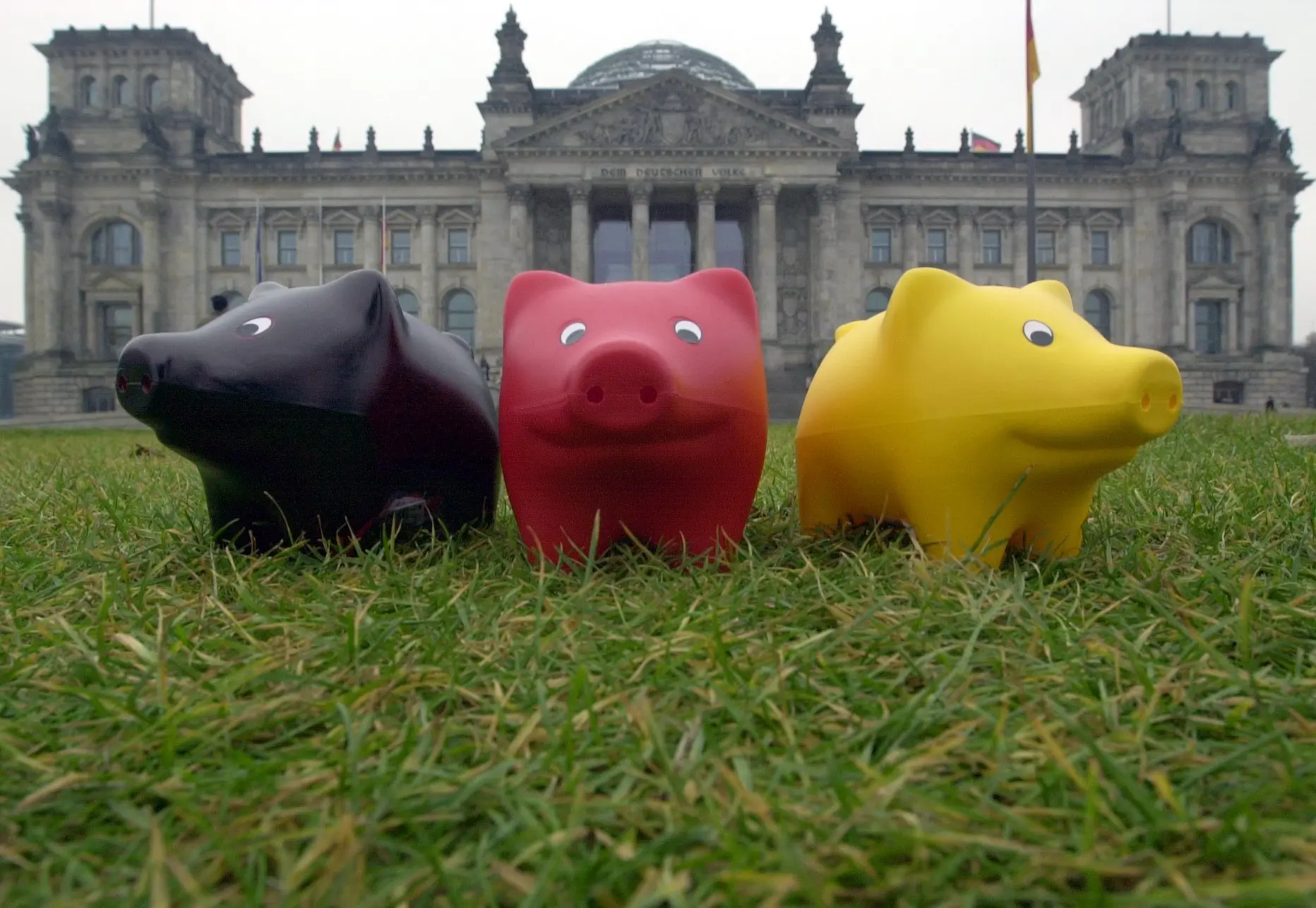 Drei Schweinchenfiguren (schwarz, rot und gold) stehen im Gras vor dem Reichstagsgebäude