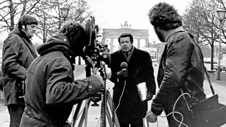 Der Journalist Lothar Loewe steht mit Mikrofon vor dem Brandenburger Tor.