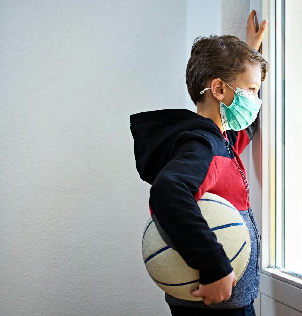 Ein Kind mit Maske und Ball unter dem Arm schaut traurig aus dem Fenster