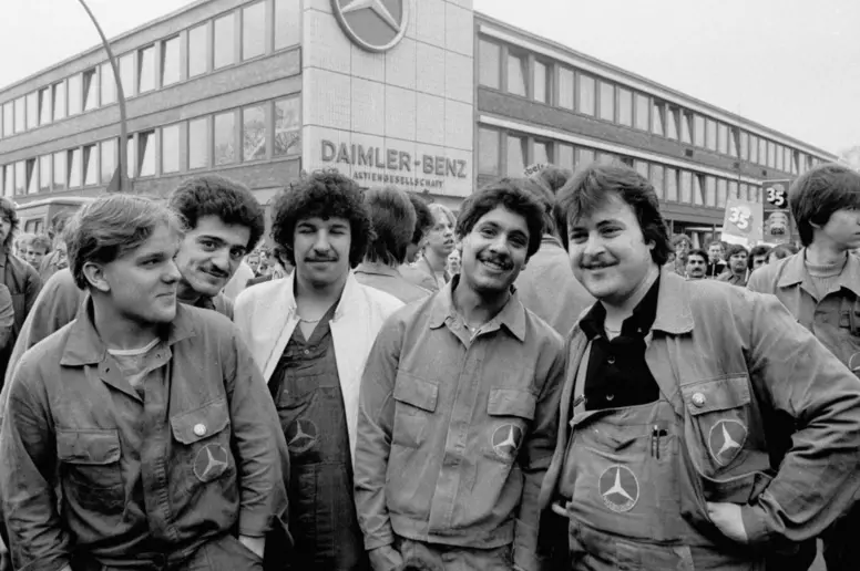Fünf türkische Gastarbeiter im Jahr 1979 stehen in Arbeitskleidung vor dem Gebäude Daimler-Benz und lächeln