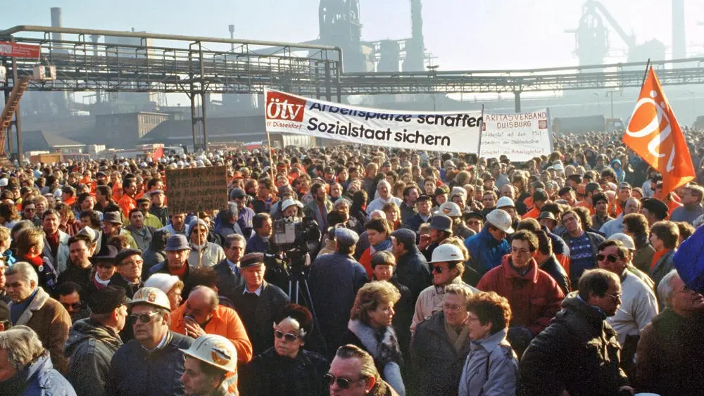 Demonstranten versammeln sich, Stahlwerk im Hintergrund.