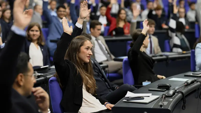 Junge Erwachsene von "Jugend und Parlament" sitzen im Plenarsaal und heben die Hände.