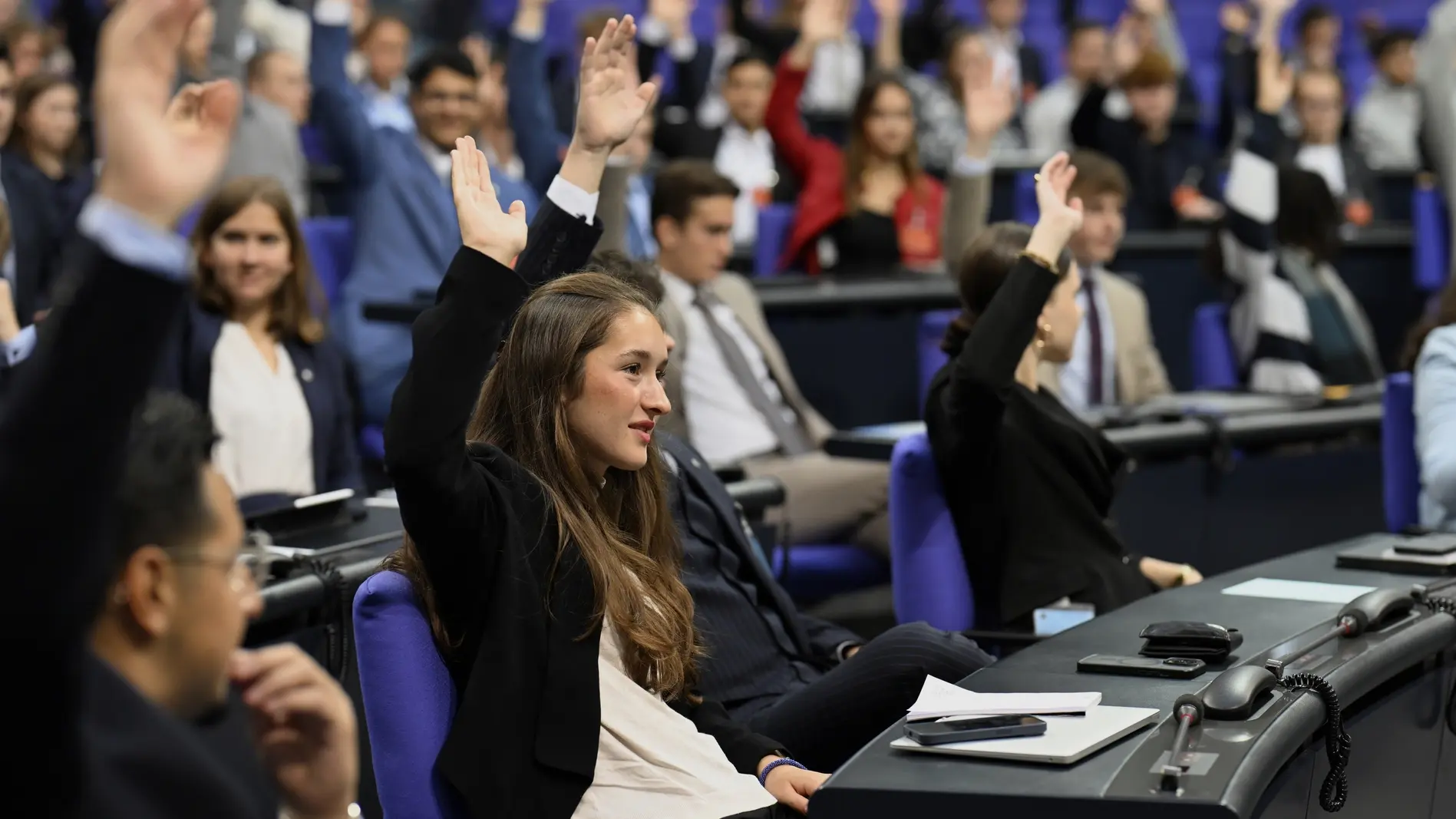 Jugend und Parlament Junge Erwachsene von "Jugend und Parlament" sitzen im Plenarsaal und heben die Hände.