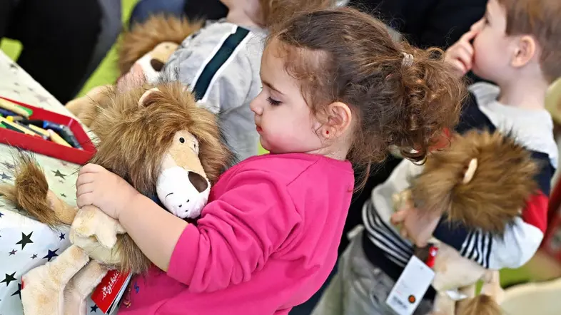 Kleinkind im pinken Oberteil steht an einem Kindertisch und hält ein Kuscheltier