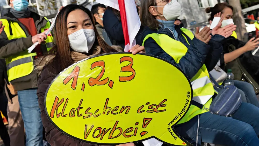 Eine Frau trägt eine medizinische Maske und hält ein Plakat "1, 2, 3 Klatschen ist vorbei!" in die Kamera.