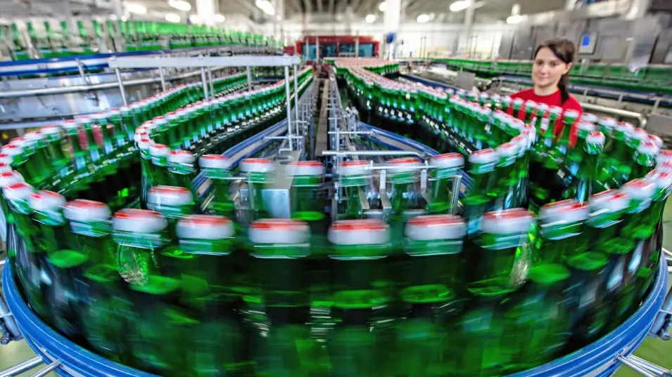 Fabrik in der grüne Glasflaschen auf einem Fabrikband am Fotograf vorbei ziehen, eine Mitarbeiterin im roten Shirt im Hintergrund.