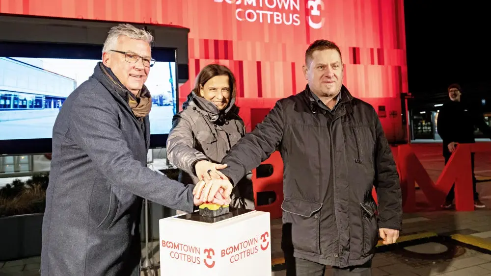 Zwei Männer und eine Frau in Winterkleidung drücken auf einen Buzzer.