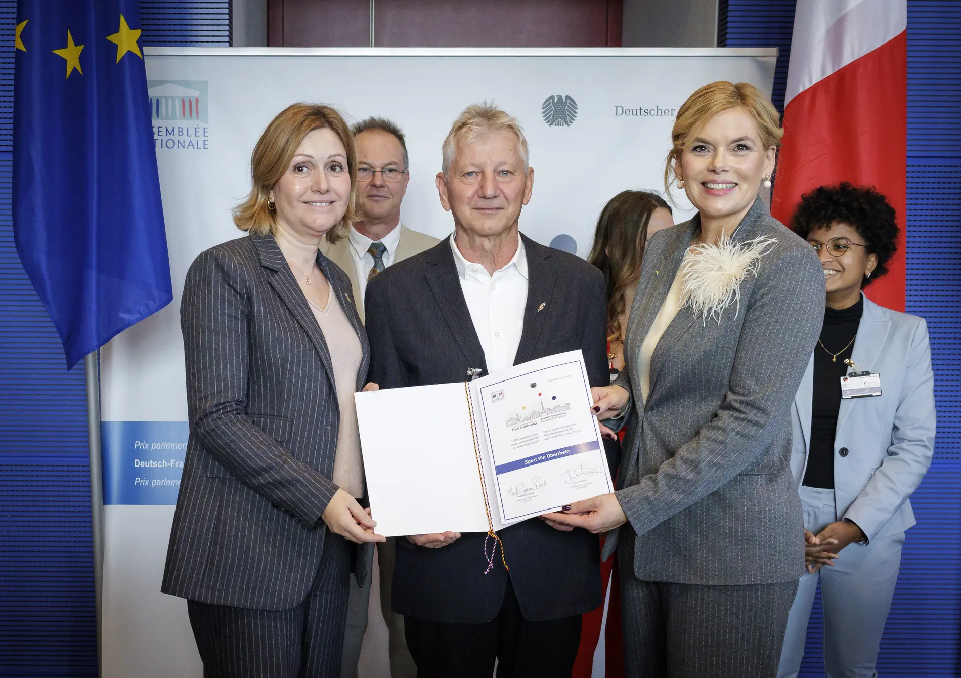 Julia Klöckner und Yaël Braun-Pivet übergeben einem Mann eine Urkunde. 