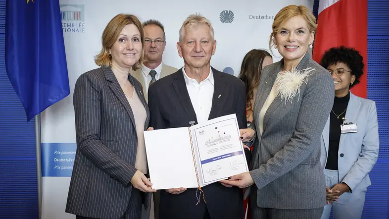 Julia Klöckner und Yaël Braun-Pivet bei der Übergabe der Urkunde an Preisträger Joachim Halm vom Projekt „Sport Pin Oberrhein“.