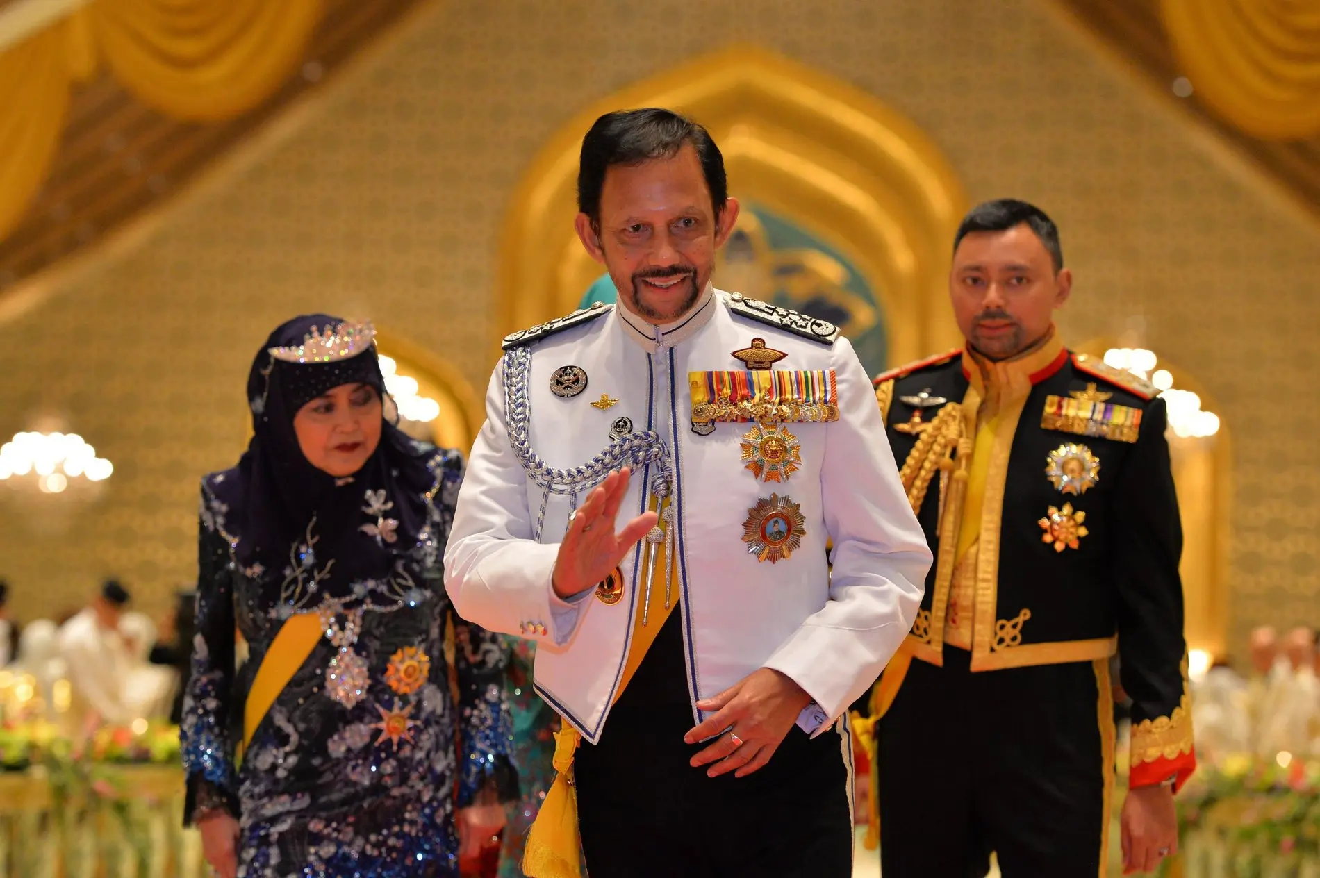 Zu sehen ist  der Sultan von Brunei zusammen mit einem Mann und einer Frau. Er hat die Hand zum Gruß erhoben und lächelt.