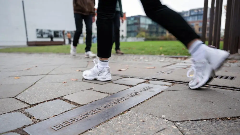 Gedenkstätte Berliner Mauer Eine Person geht an der Gedenkstätte Berliner Mauer in der Bernauer Straße über einen Streifen am Boden, der mit "Berliner Mauer" gekennzeichnet ist und den ehemaligen Verlauf nachzeichnet.